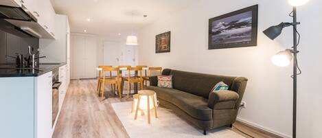 Une cuisine moderne avec des meubles blancs et un plan de travail noir, une table à manger en bois avec des chaises, un canapé gris agrémenté d’un coussin décoratif et un lampadaire.