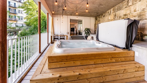 Un jacuzzi de madera en un balcón con vistas a zonas verdes y edificios.