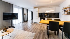 Una sala de estar moderna con un televisor de pantalla plana, una mesa de comedor de madera y una cocina con armarios blancos y encimera negra.