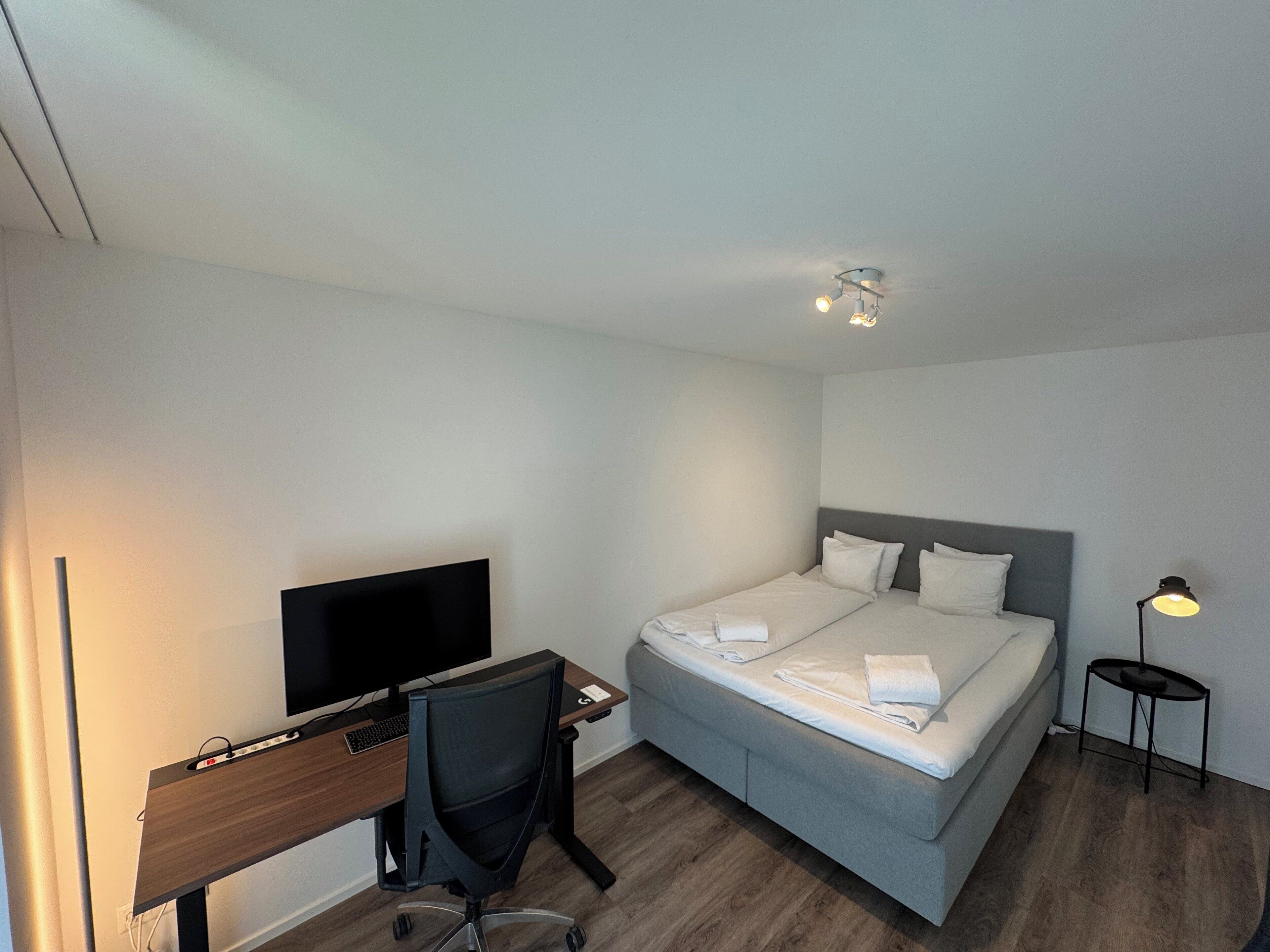 A hotel room with a bed, a desk with a computer, a chair, and a bedside table with a lamp.