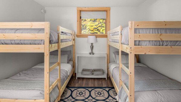 Una habitación pequeña con dos literas, una mesita con lámpara, y una ventana que da a vista de árboles.