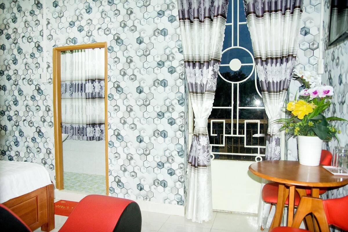 Habitación de hotel con pared de diseño hexagonal, cama de madera, sofá rojo, mesa de comedor con un jarrón de flores y cortinas con diseño geométrico.