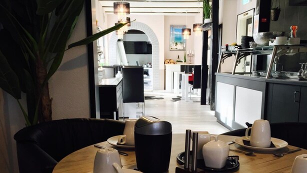 Zona de menjador amb una taula rodona preparada per a dinar, un espai per al cafè i vista cap a la cuina.