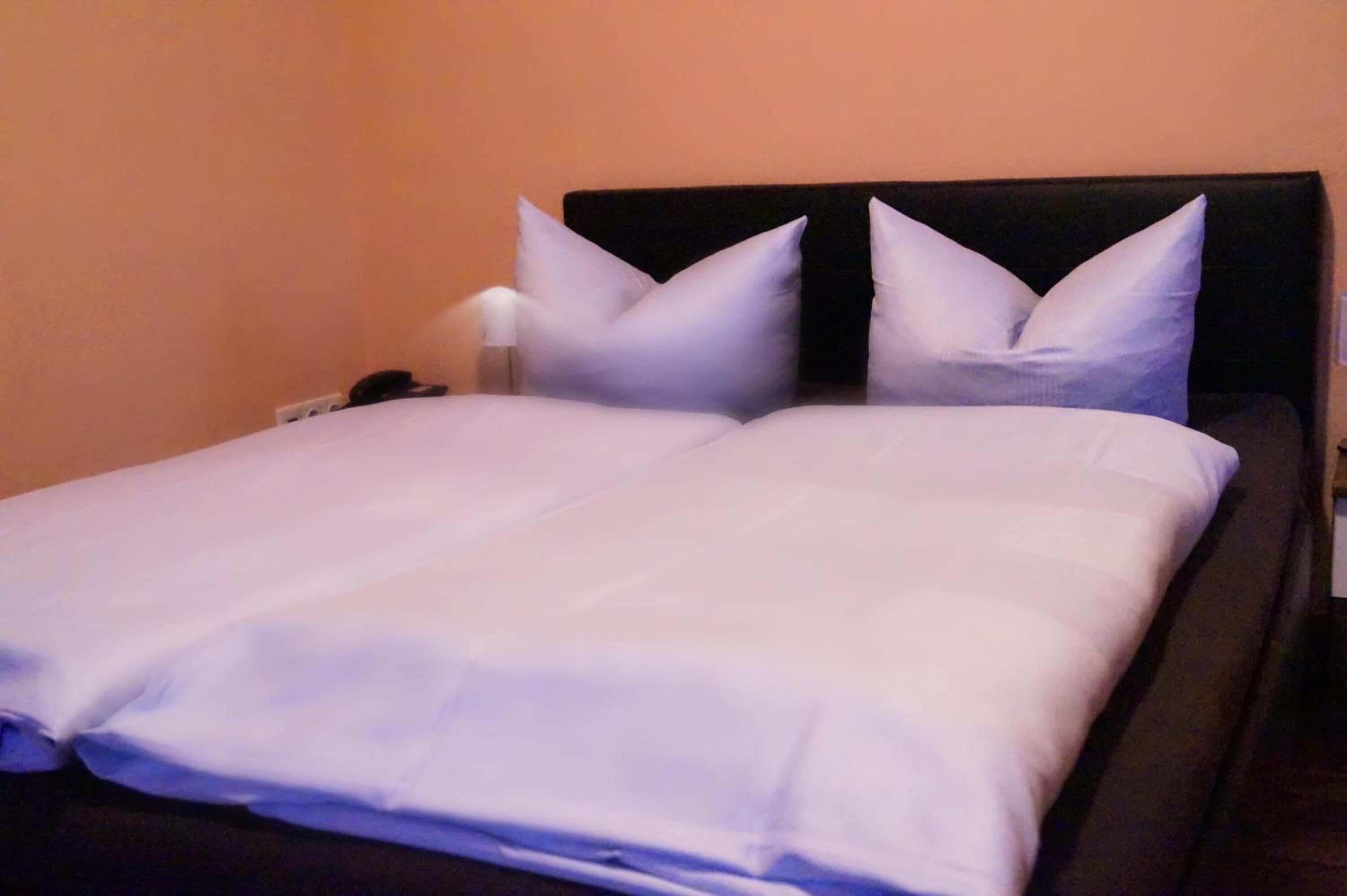 A neatly made bed with white linens and two pillows, a bedside table with a lamp, and a wall-mounted phone.