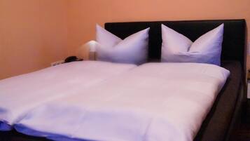 A neatly made bed with white linens and two pillows, a bedside table with a lamp, and a wall-mounted phone.