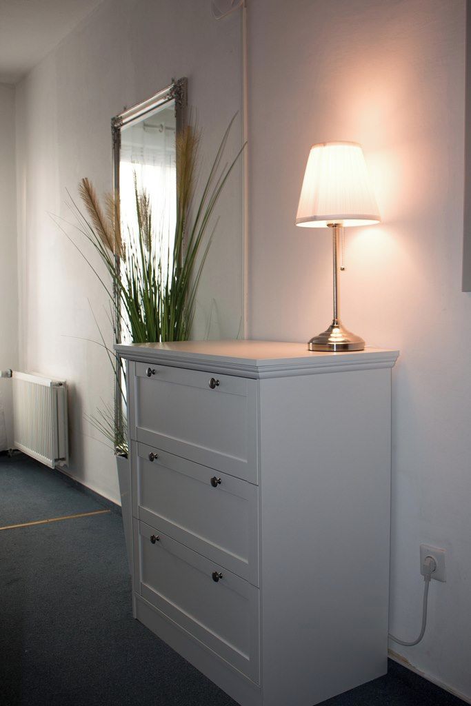 Ein weißer Kommode mit vier Schubladen, ein Spiegel darüber und eine Lampe obenauf.