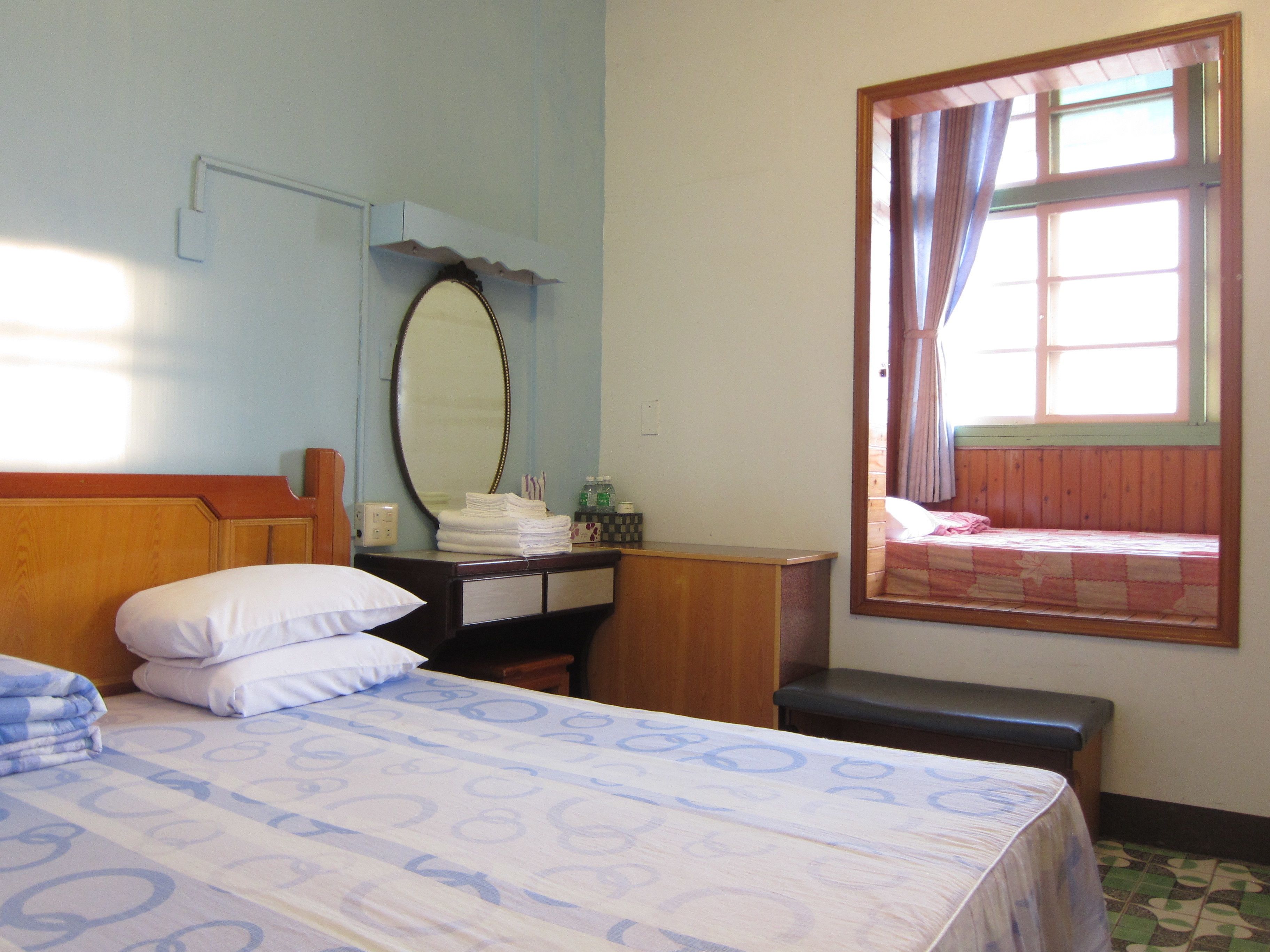 A hotel room with a bed, a wooden headboard, a bedside table with a mirror, a window with curtains, and a small bench.