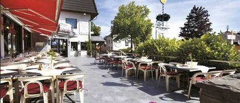 Un café al aire libre con mesas y sillas, un toldo rojo y un edificio con un letrero al fondo.