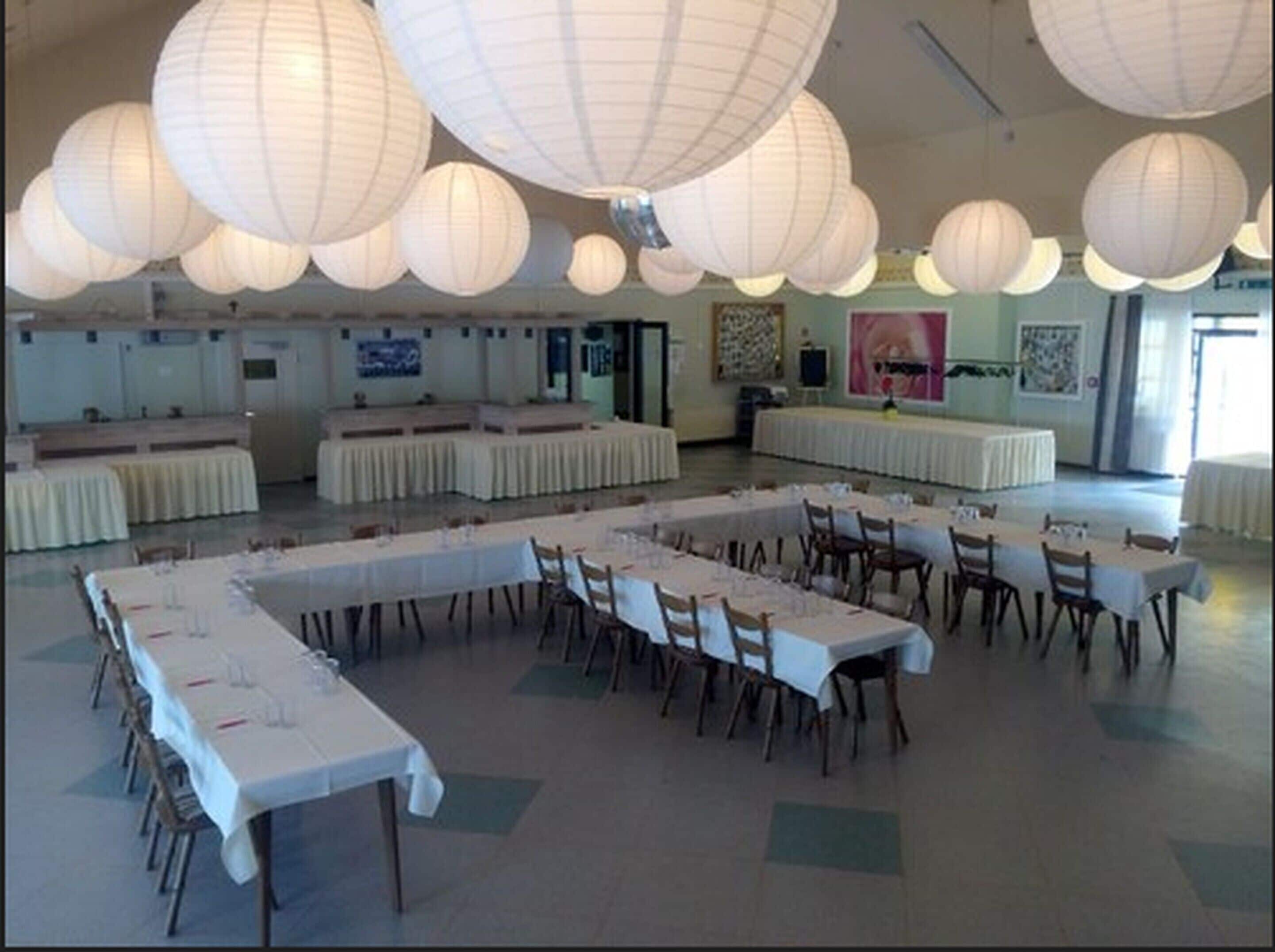 A banquet hall with long tables set for dining, white paper lanterns hanging from the ceiling, and a stage area in the background.