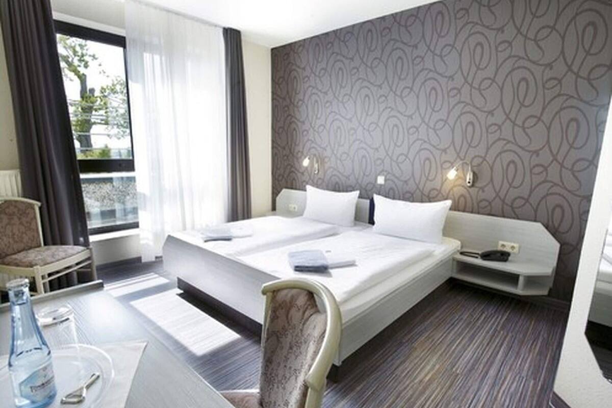 A hotel room with a large bed, a desk with a chair, a small table with a bottle and spoon, and a view of the outdoors.