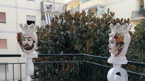 A balcony with a white pot of red flowers, a metal railing, and a building in the background. - Al Parco Primavera (Sorrento)