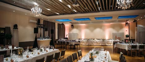 A spacious banquet hall with long tables set for dining, wooden ceiling panels, and chandeliers.