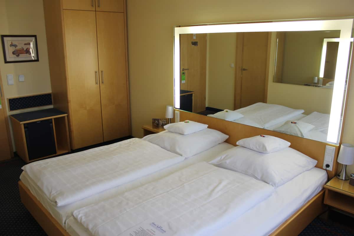 A hotel room with two single beds, a large mirror, a bedside table with a lamp, and a small desk with a chair.