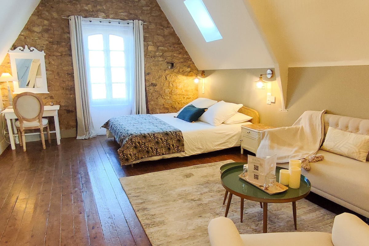 A cozy bedroom with a sloped ceiling, a bed with a patterned bedspread, a bedside table with a lamp, a chair, and a small table with candles and a tray.