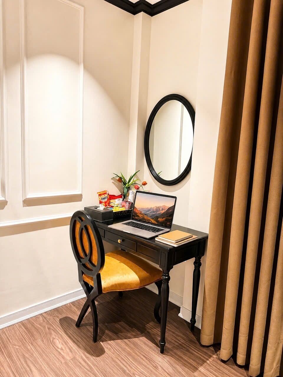 Un escritorio con una computadora portátil, un cuaderno y un espejo en una habitación con ventana y cortinas.