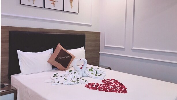 Ein Hotelzimmer mit einem Bett, weißer Bettwäsche, einem Kopfkissen mit Hotel-Logo, einem weißen Telefon und einer herzenförmigen Anordnung aus roten Rosenblättern.