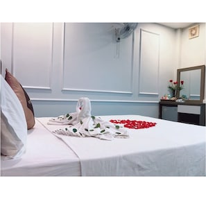A hotel room with a neatly made bed, a towel with a heart, and red rose petals on the bed.