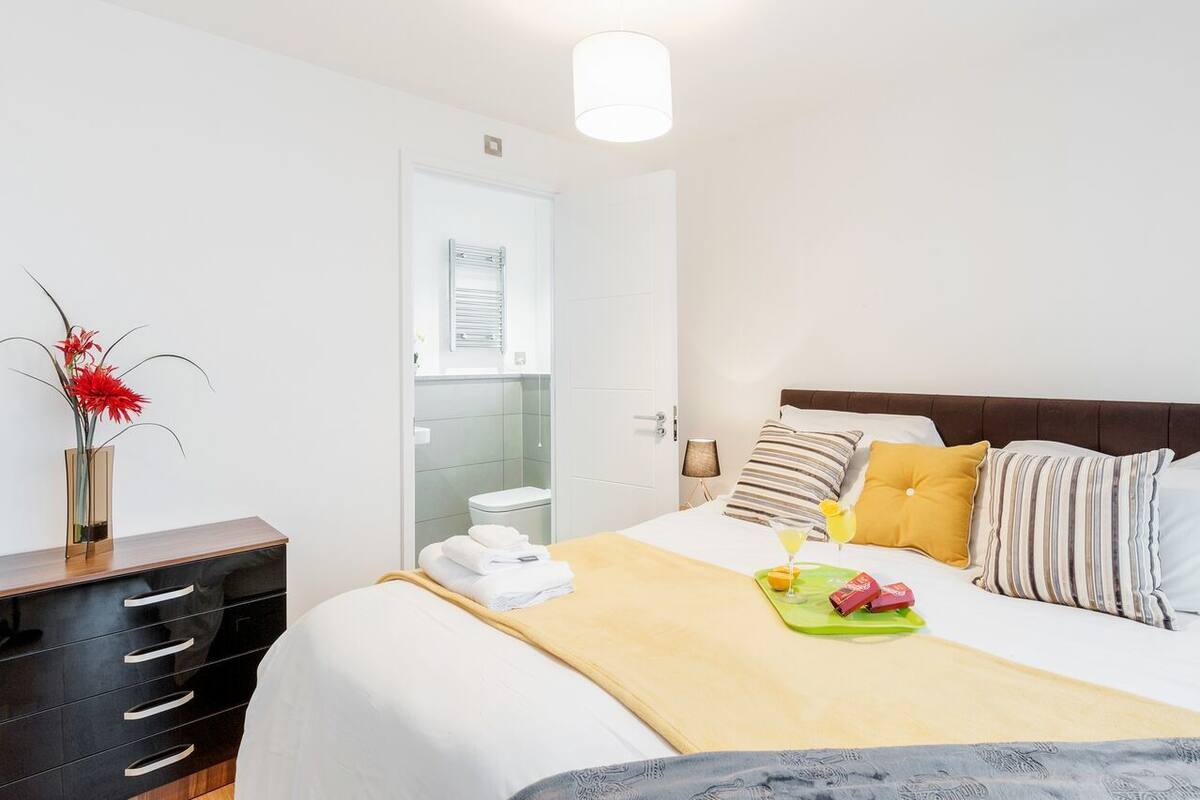 Une chambre d’hôtel avec un lit, une table de chevet, une lampe, un vase de fleurs et une salle de bain visible à travers une porte ouverte.
