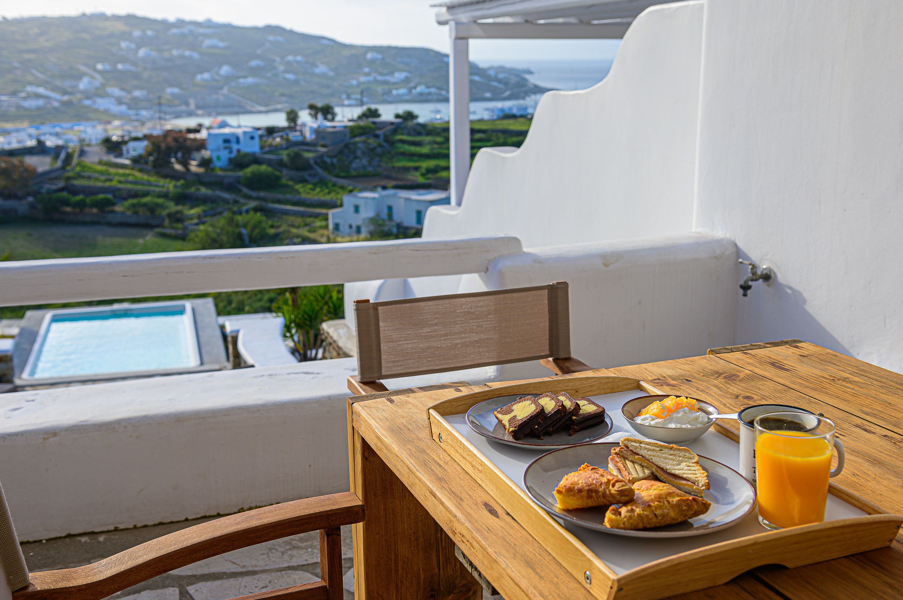 A wooden table with breakfast items including croissants, a plate of bread, a bowl of yogurt, and a glass of orange juice, set against a white wall with a view of a coastal landscape.
