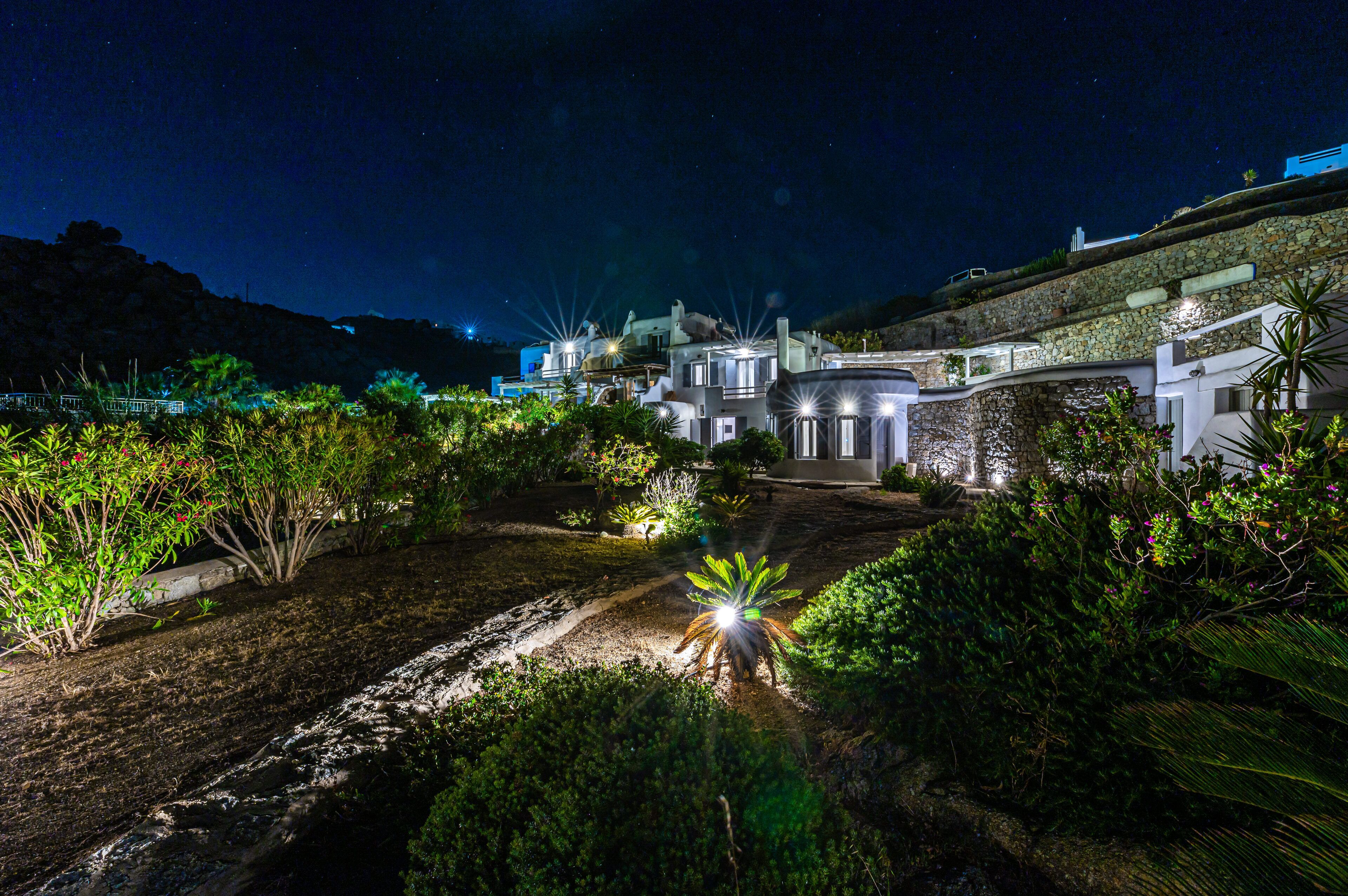 A nighttime view of a coastal property with stone walls, a well-lit garden, and a clear view of the sea.