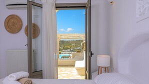 A hotel room with a bed, a bedside lamp, and a view of a pool and buildings. - 9 Muses Villas Mykonos (Mykonos)