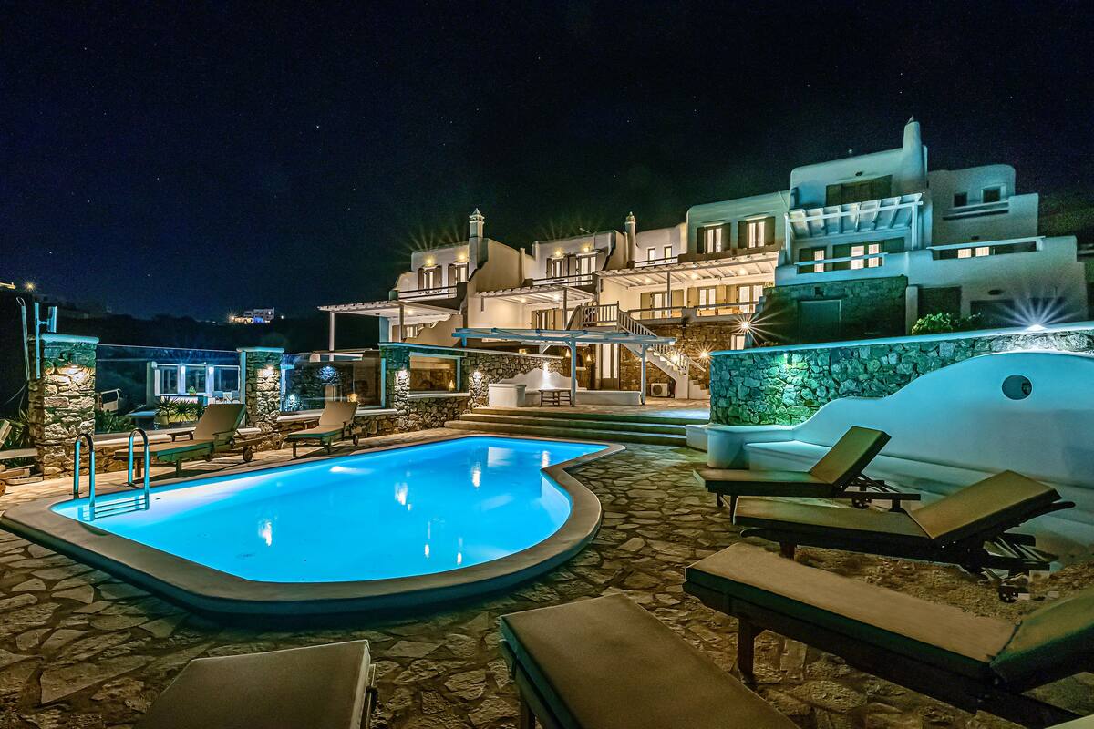 A nighttime swimming pool area with lounge chairs, a stone-tiled deck, and a modern building with multiple balconies and lights.