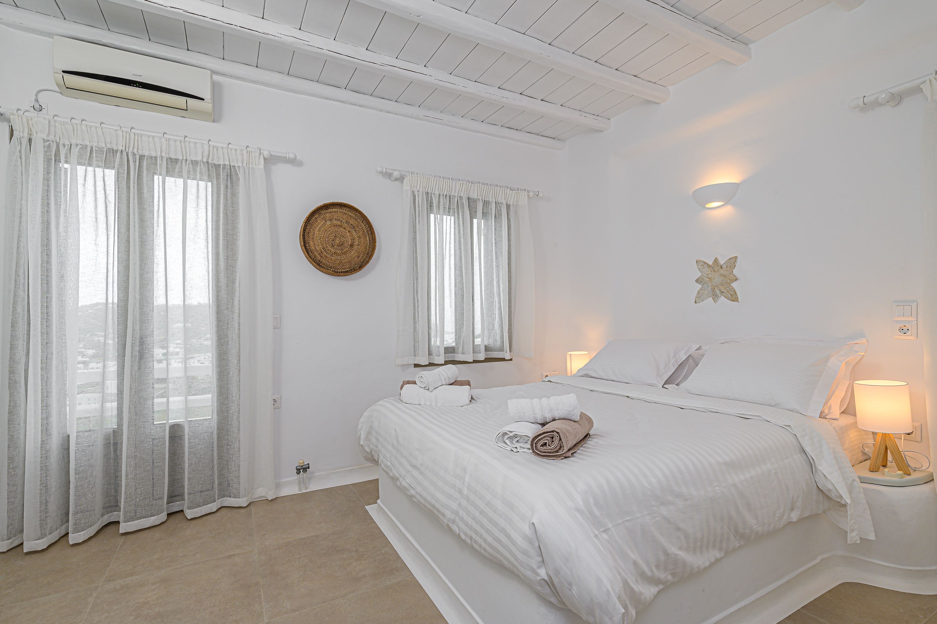 A white bedroom with a bed, curtains, a wall-mounted air conditioner, a bedside lamp, and a decorative wall piece.