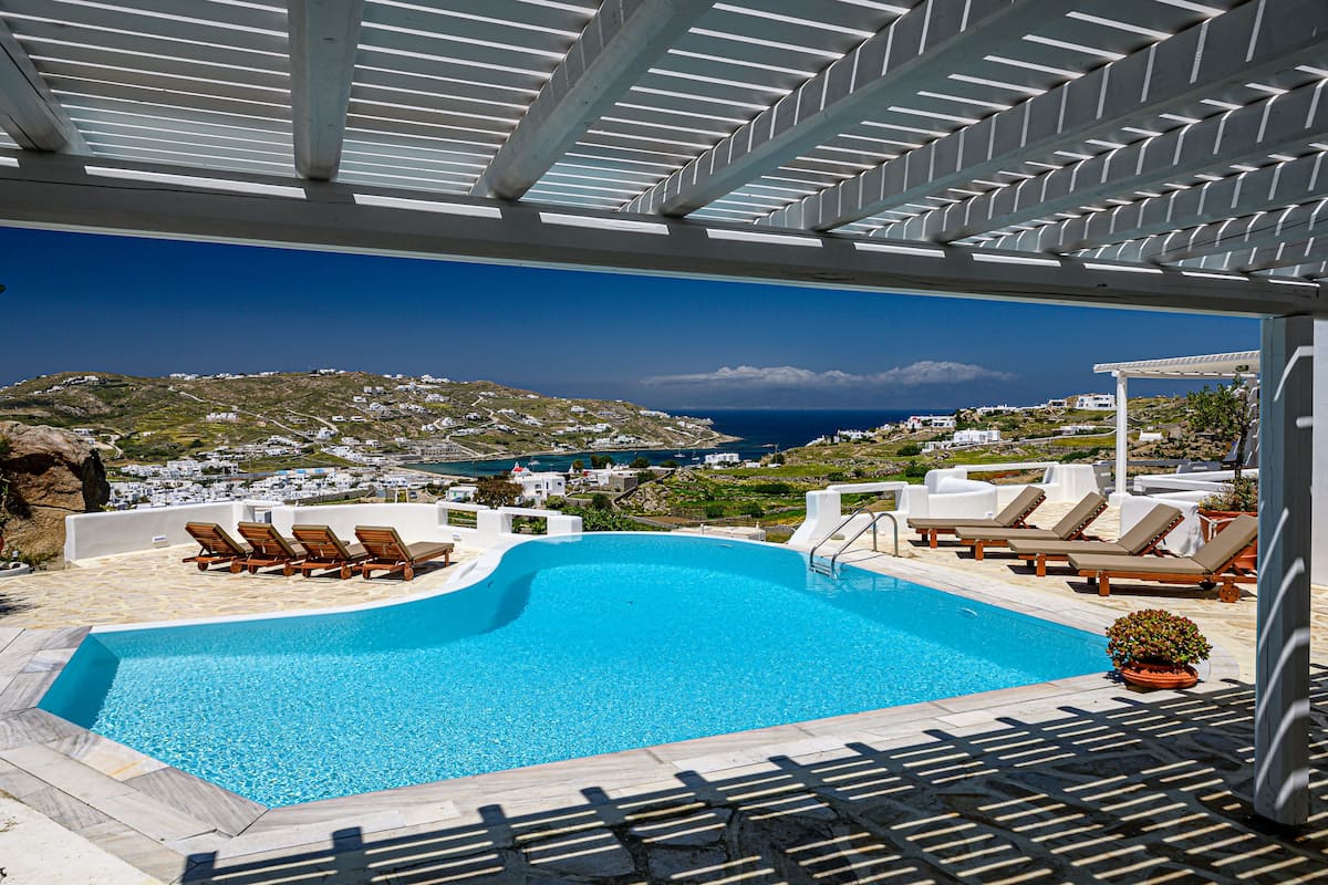 A pool with lounge chairs, a pergola, and a view of a coastal town.