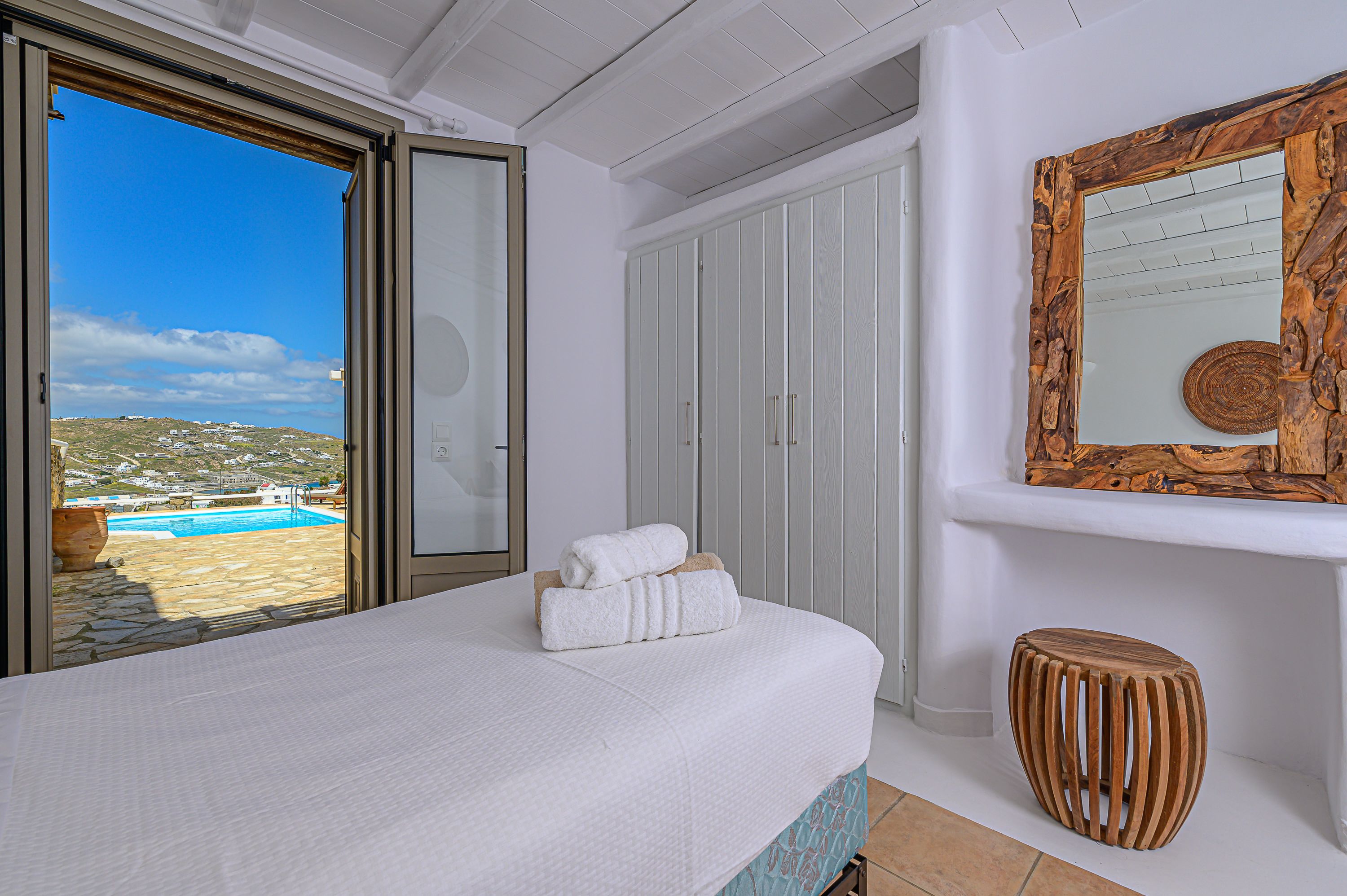 A bedroom with a large bed, a wooden stool, a wooden-framed mirror, and a view of a pool and landscape.
