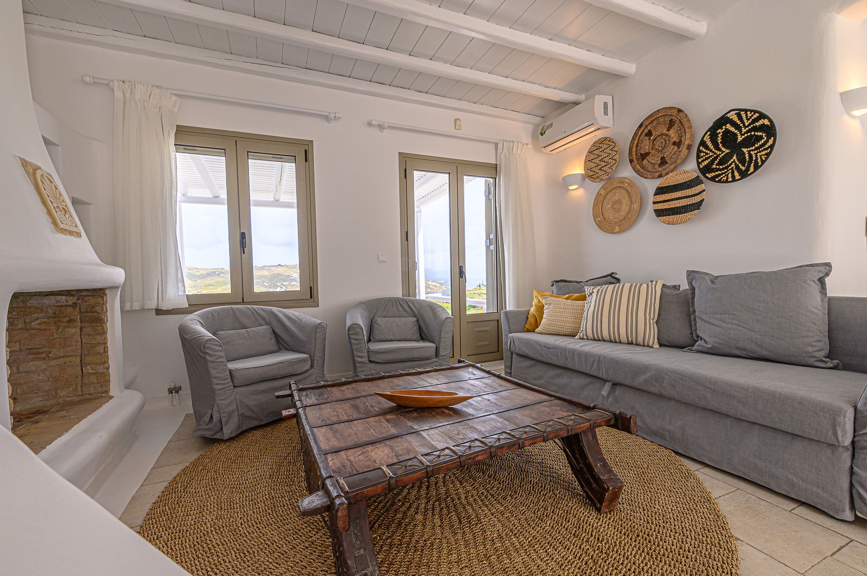 A living room with a fireplace, grey sofas, a wooden coffee table, and a view through large windows.