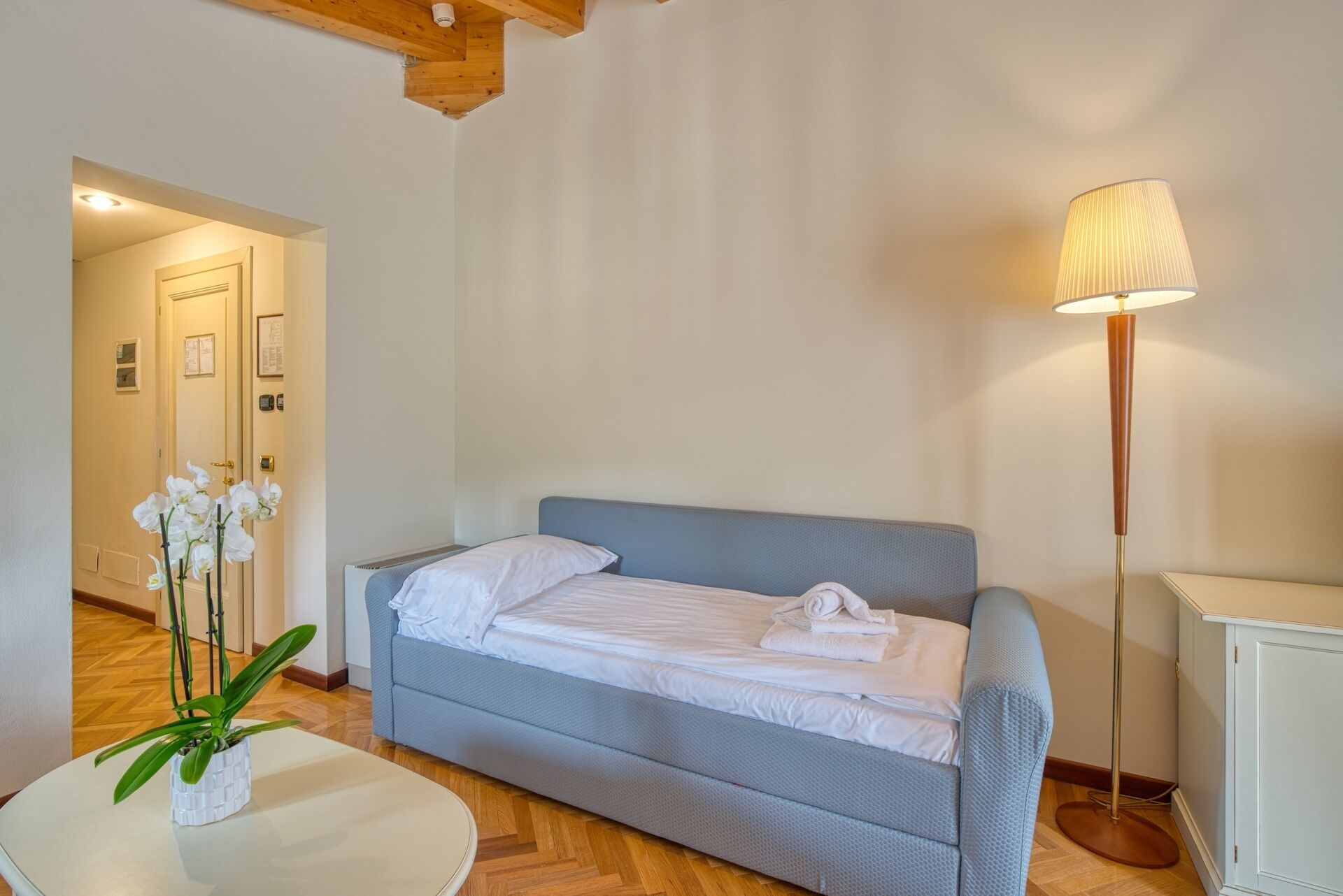 Ein Schlafzimmer mit einem Bett, einer Stehlampe, einem weißen Tisch mit Pflanze und einer Tür zu einem anderen Raum.