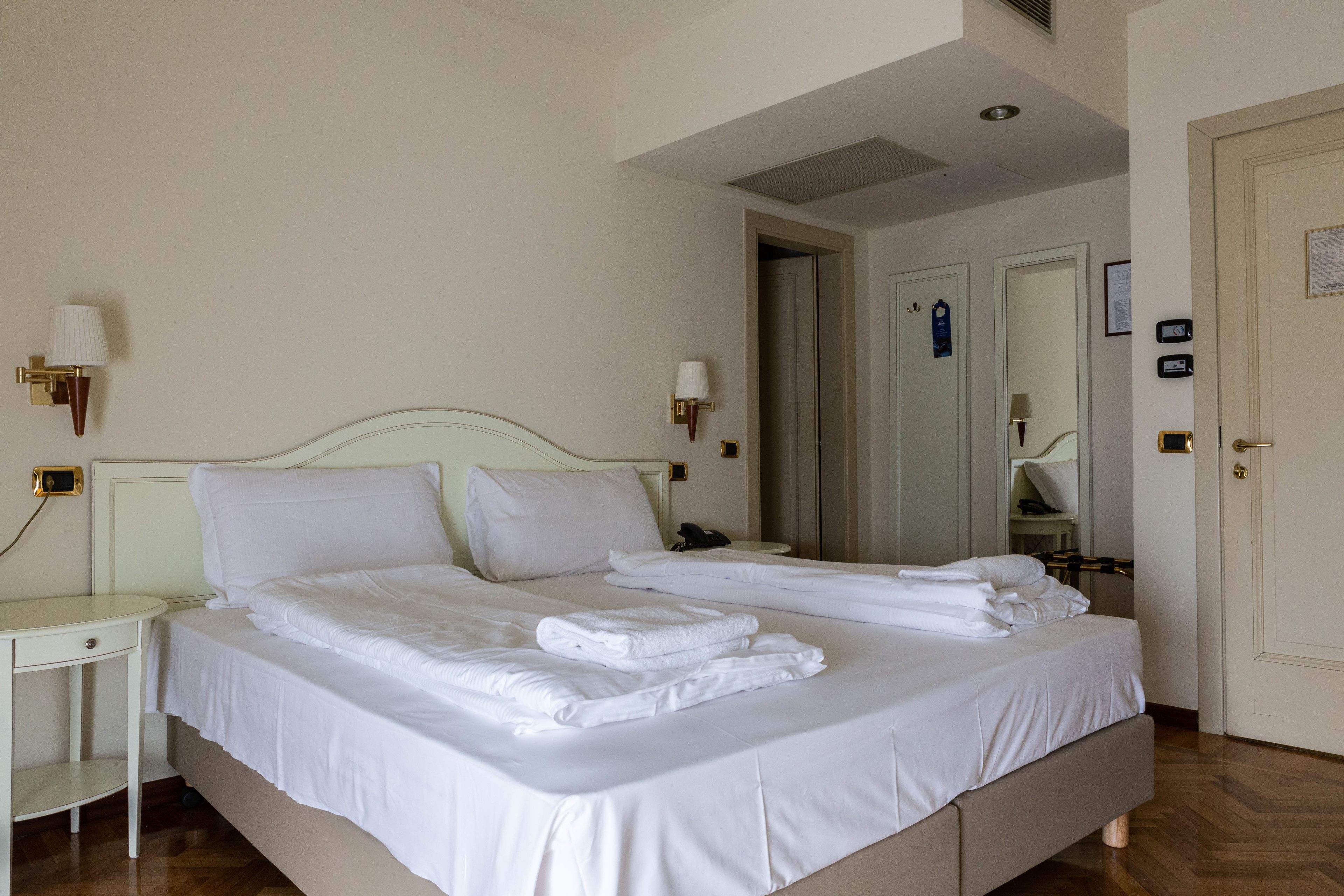 A neatly made bed with white linens and towels, a bedside table with a lamp, and a door with a mirror and a coat rack.