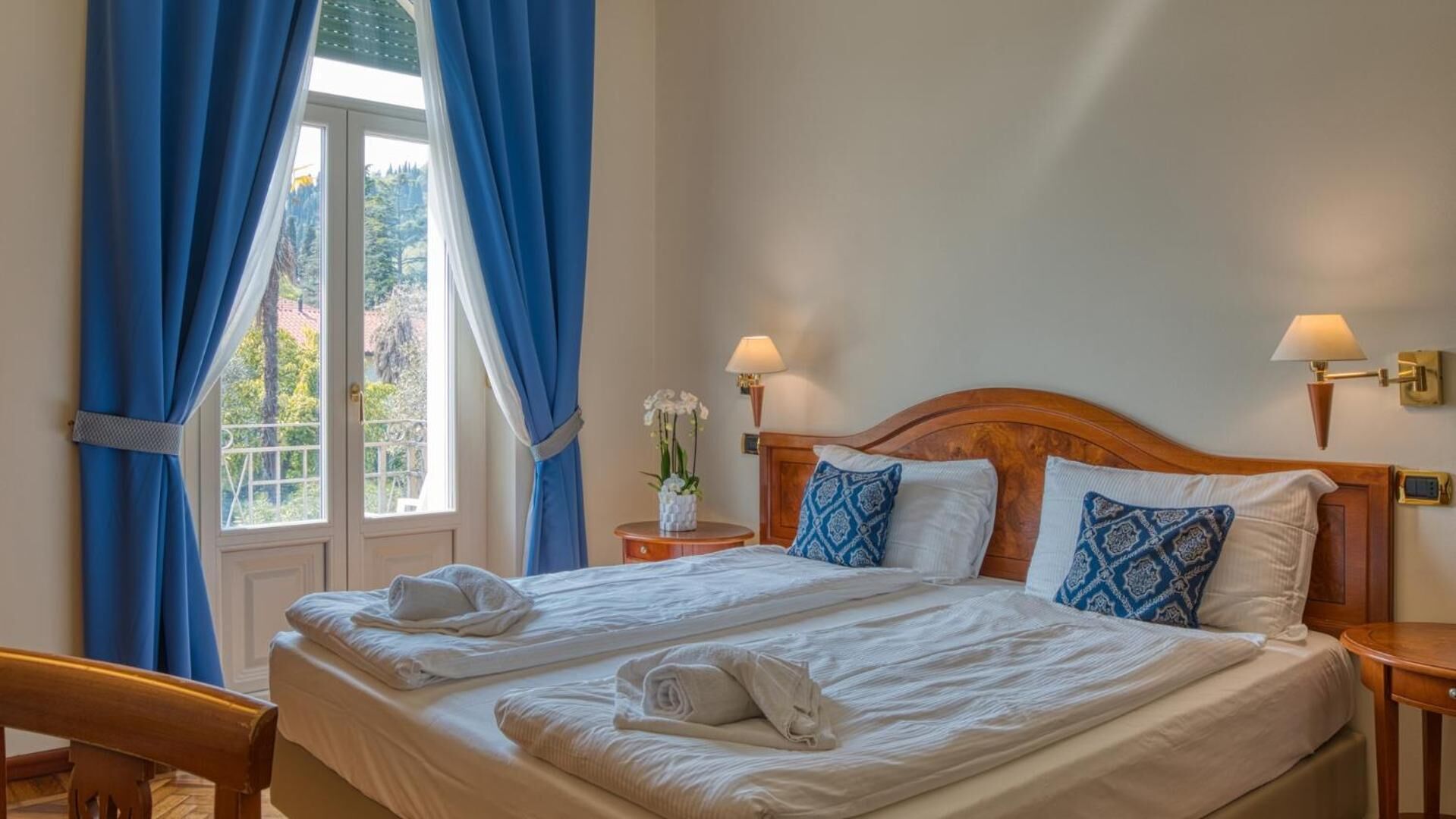 A double bed with white linens, two patterned pillows, a wooden headboard, and a nightstand with a lamp.