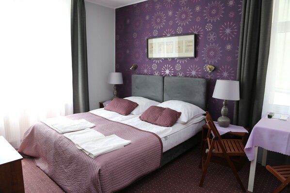 Un lit bien fait, recouvert d’une couvre-lit violette et blanche, avec deux oreillers et une table de chevet surmontée d’une lampe.