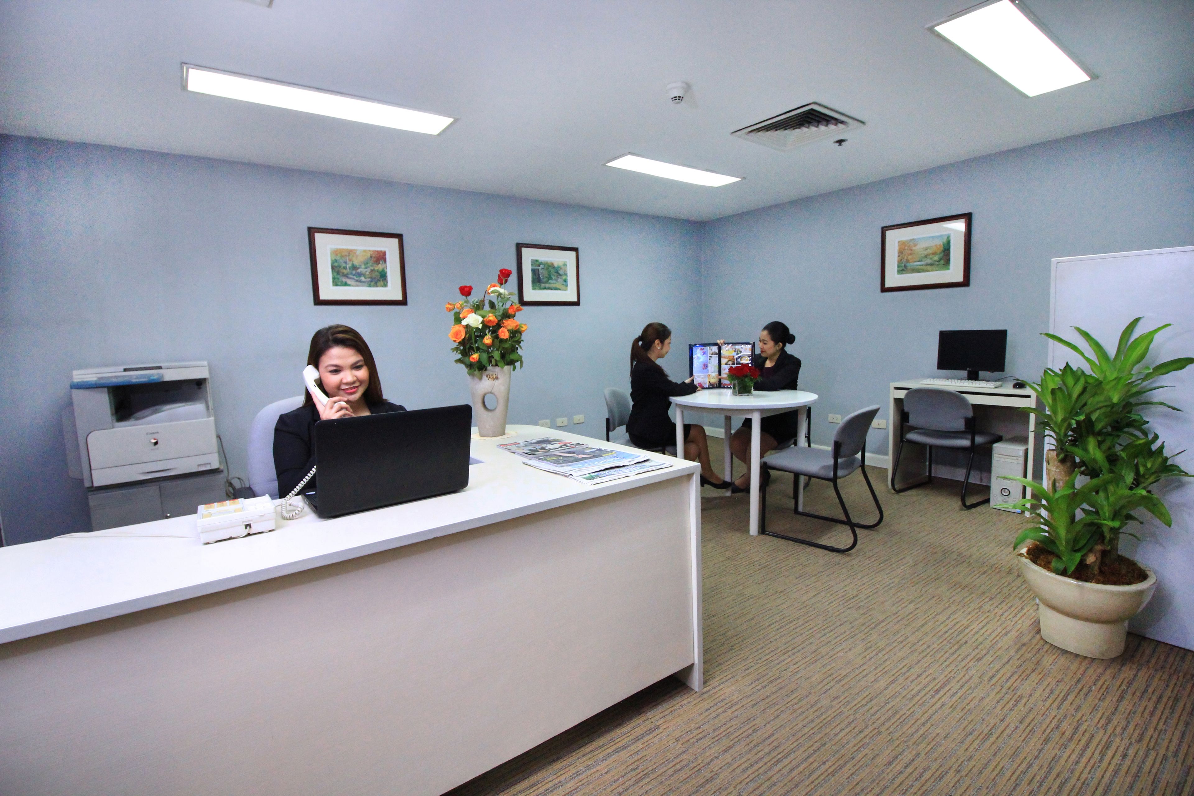 An office with a reception desk, computers, and chairs. There are framed pictures on the walls and a potted plant.