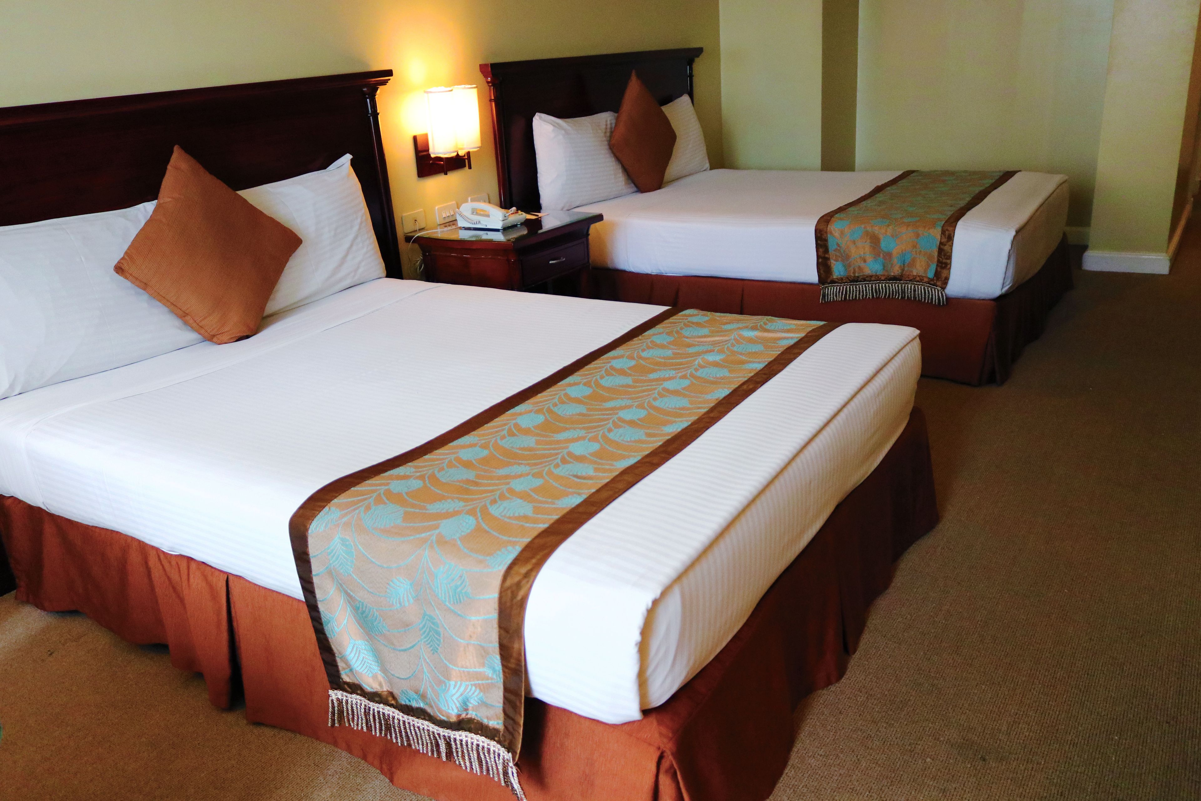 Duas camas de casal com cabeceiras marrons, lençóis brancos e almofadas marrons. Cada cama tem uma mesa de cabeceira de madeira com uma luminária e um telefone.