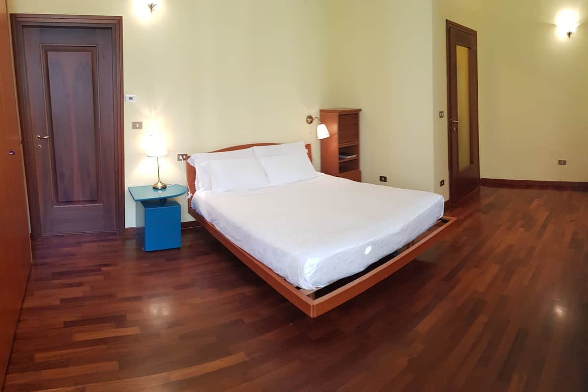 A bedroom with a wooden bed, a nightstand with a lamp, a blue dresser, and a wooden wardrobe.