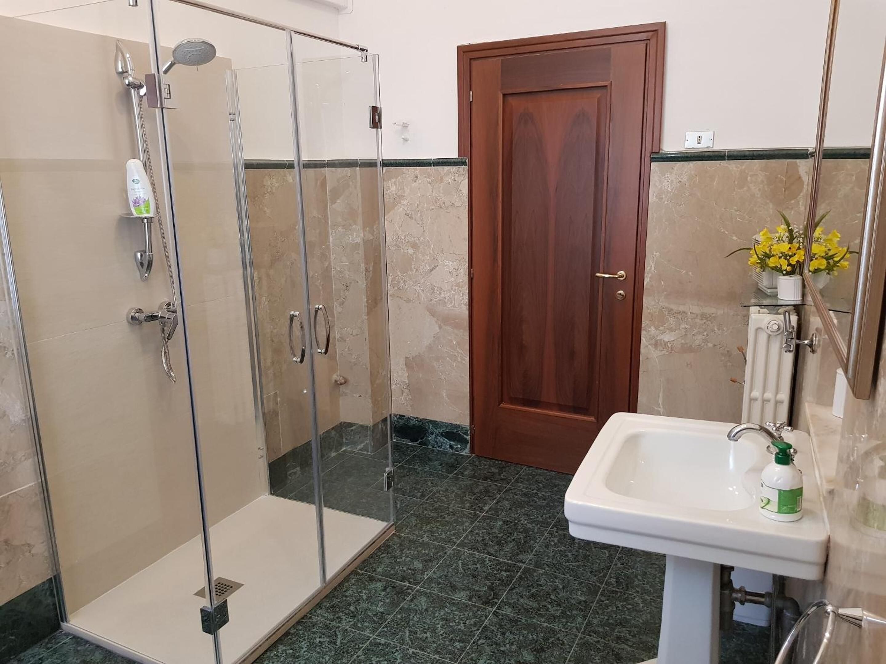 Un bany amb dutxa envidriada, una porta de fusta, un lavabo amb mirall i un ram de flors.