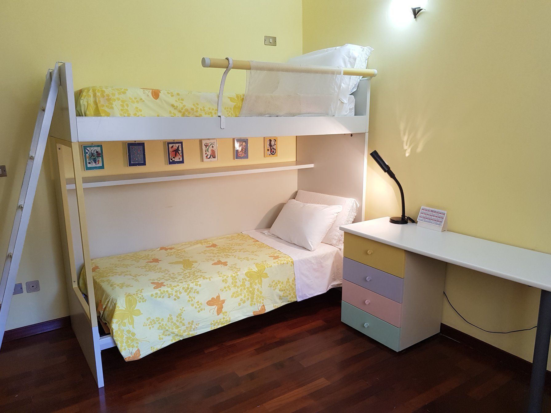 A bunk bed with a desk and drawers, a ladder, and a wall-mounted shelf with pictures.