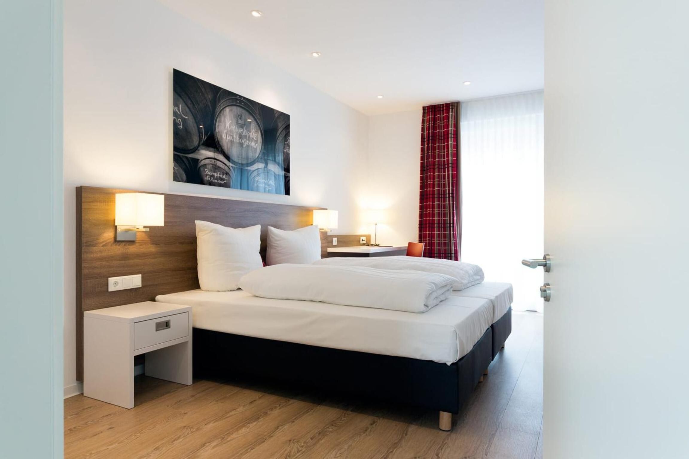 A modern hotel room with a large bed, bedside tables, a wall-mounted artwork, and a window with curtains.