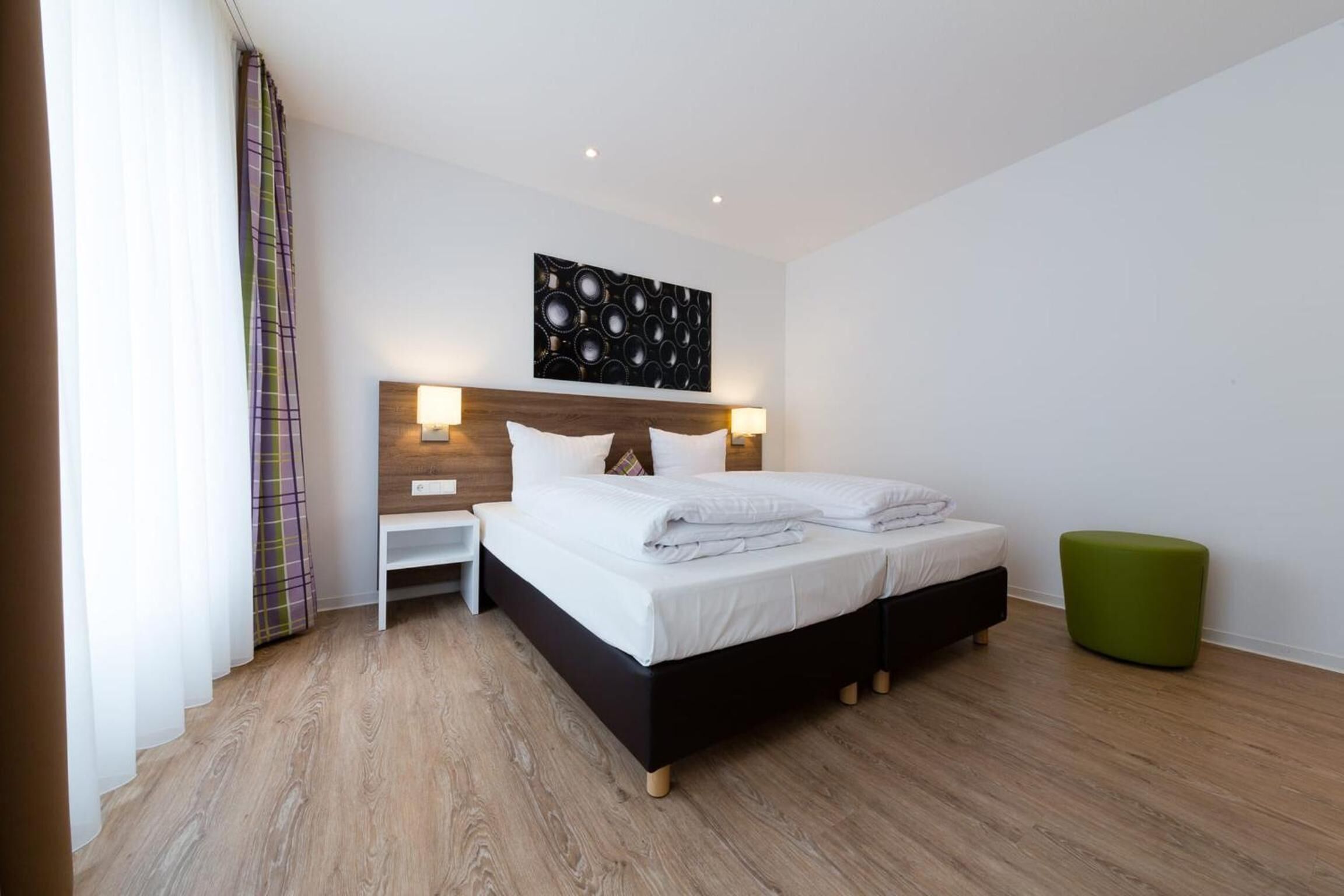 Quarto de hotel moderno com uma cama grande, mesas de cabeceira, um banquinho verde e uma obra de arte pendurada na parede.