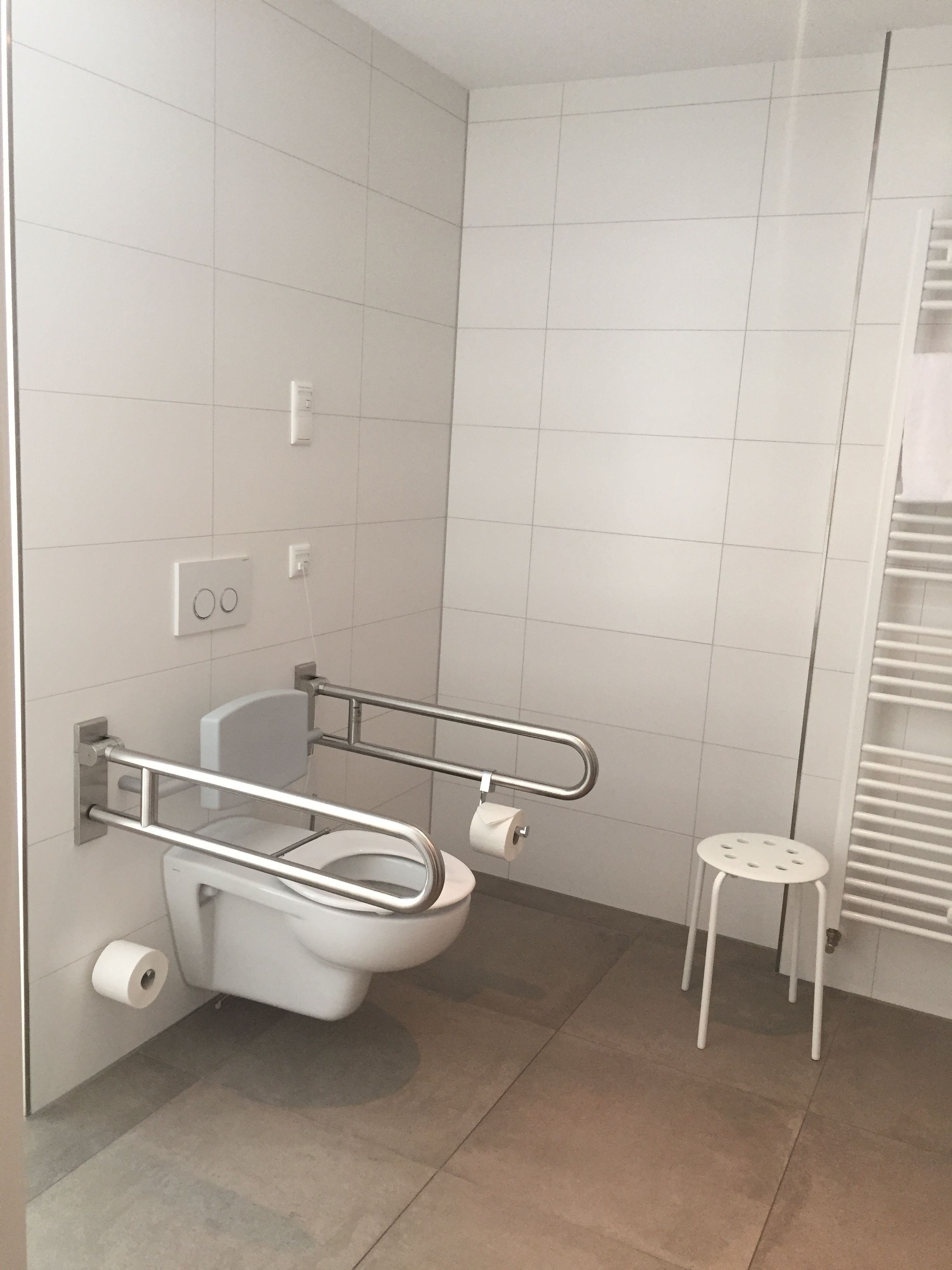 A modern bathroom with a white toilet, a handrail, a small stool, and a towel rack.