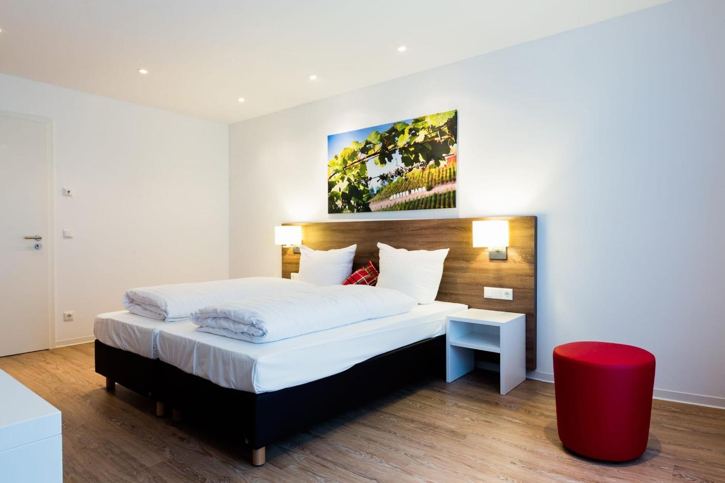 Une chambre d’hôtel moderne avec un grand lit, des tables de chevet, un pouf rouge et un tableau représentant un vignoble, accroché au mur.