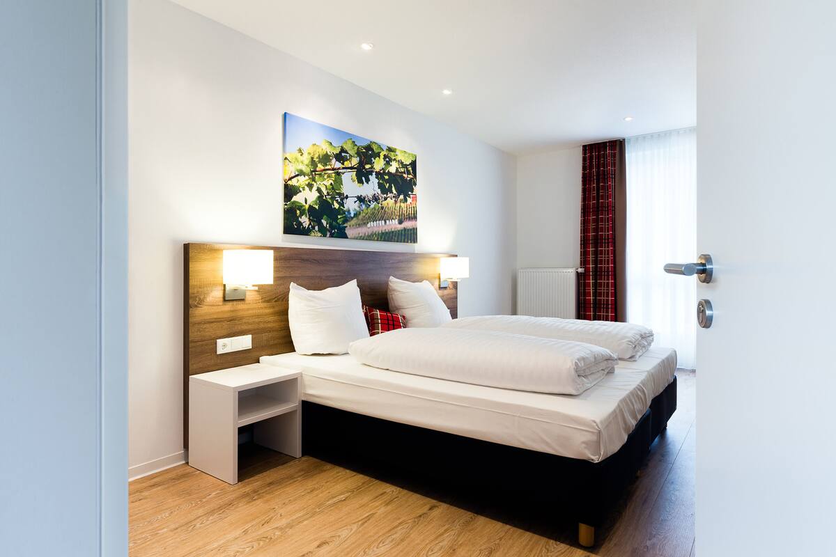 Moderni hotellihuone, jossa on suuri sänky, yöpöydät, sängynpääty, jossa on sisäänrakennettu hylly, ja seinälle kiinnitetty taideteos.