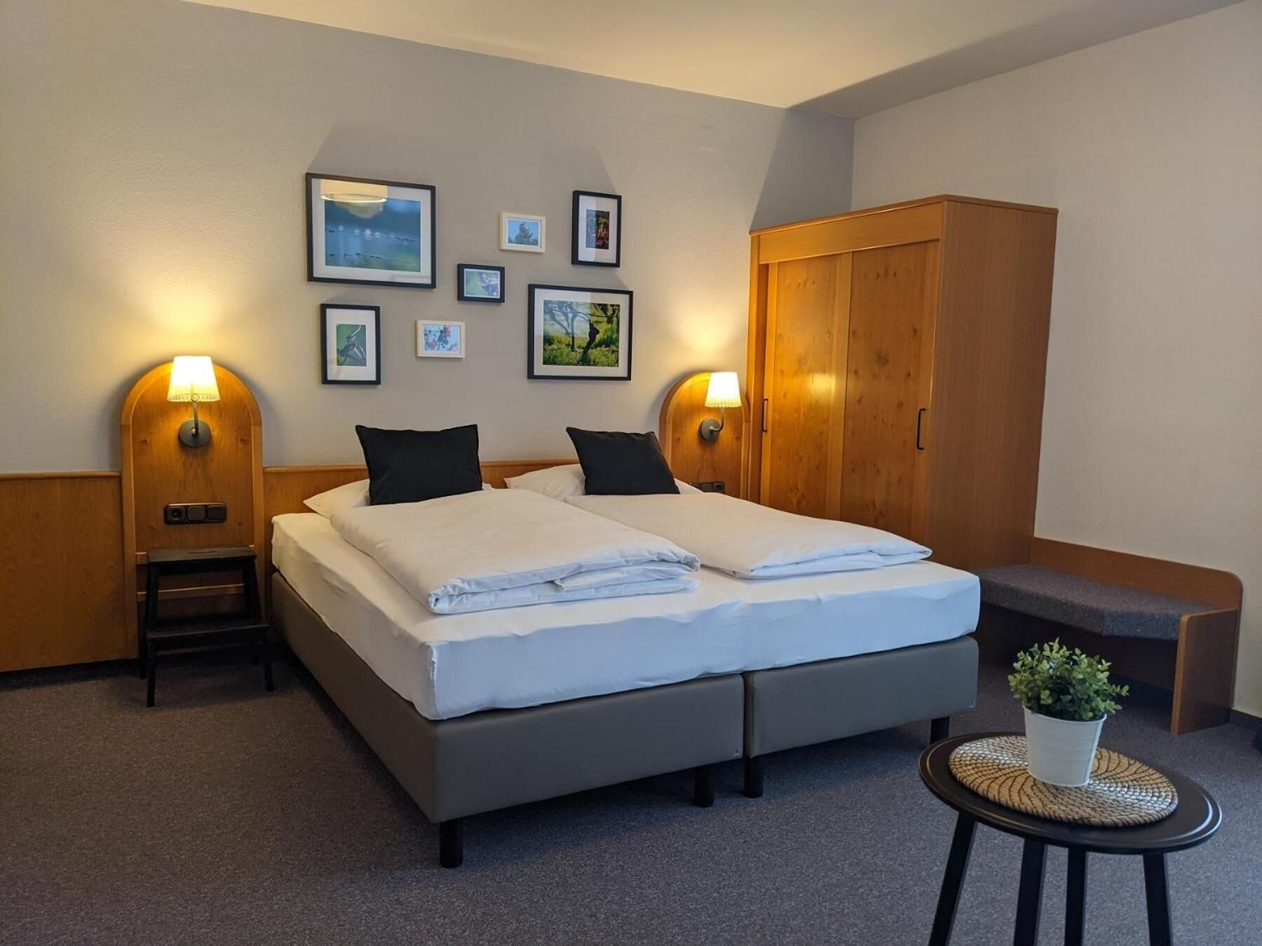 Un lit double avec du linge de lit blanc, deux tables de chevet avec des lampes, une armoire en bois et une petite table ronde avec une plante.