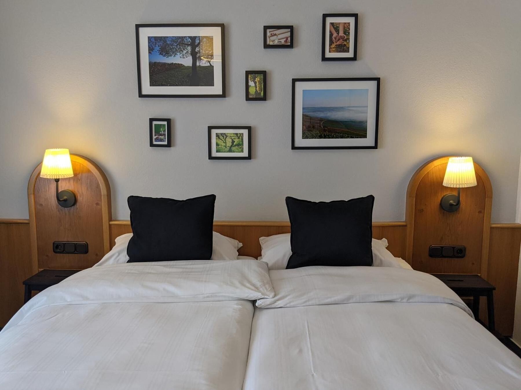 Uma cama de casal com roupa de cama branca, duas almofadas pretas e duas mesas de cabeceira com abajures. Há quadros emoldurados na parede acima da cama.