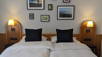 Uma cama de casal com roupa de cama branca, duas almofadas pretas e duas mesas de cabeceira com abajures. Há quadros emoldurados na parede acima da cama.