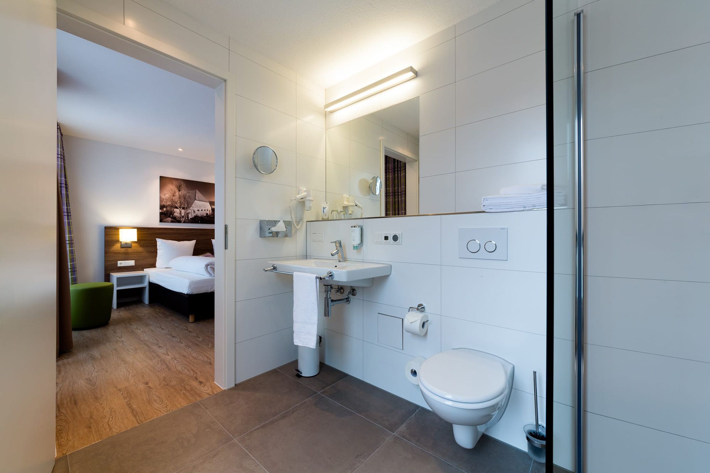 Un baño moderno con lavamanos, inodoro y espejo. Se observa una mesita de noche con lámpara y una silla verde.