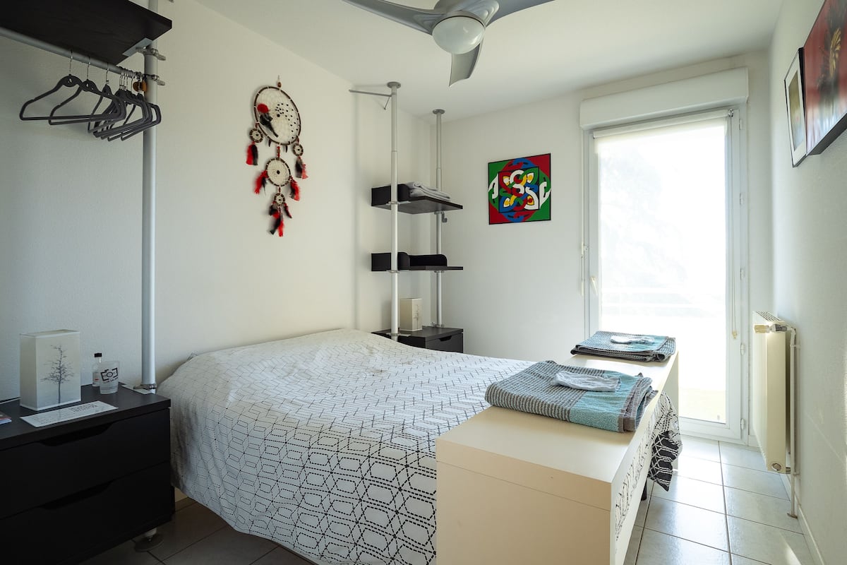Ein Schlafzimmer mit Bett, Schreibtisch, Deckenventilator, Traumfänger, farbigem Kunstwerk und Fenster.