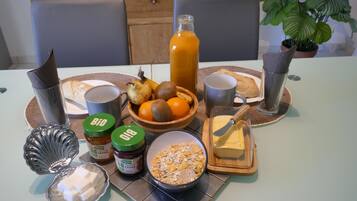 Desayuno con un bol de fruta, una botella de zumo de naranja, tostadas en un plato, un bol de cereales, un tarro de mermelada, mantequilla en un plato y un cuchillo sobre una mesa.